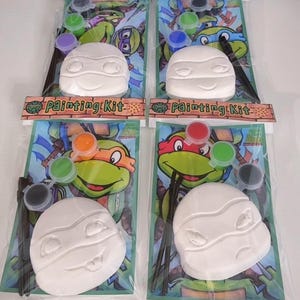 May include: Four Teenage Mutant Ninja Turtles painting kits. Each kit includes a white, unpainted turtle mask, paint pots in various colors, and a paintbrush. The background features a colorful illustration of the turtles.