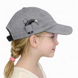 Puede incluir: Gorra de béisbol gris con un abrebotellas negro y plateado adjunto. La gorra tiene una visera curva y una correa ajustable negra. El abrebotellas es de metal y tiene un diseño curvo.