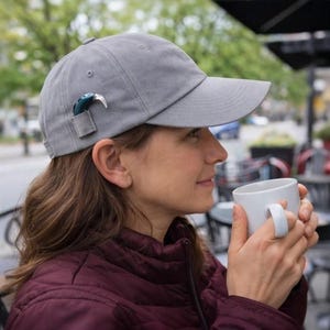 May include: A gray baseball cap with a small, dark-colored accessory attached to the side. The cap is worn outdoors. The person is holding a white mug. The background includes a street, trees, and outdoor seating.