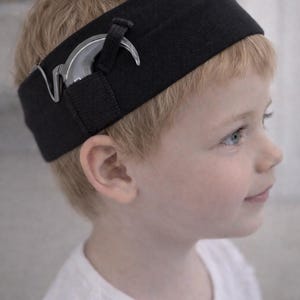 May include: A black headband with a silver accessory. The headband is worn on the head of a young child with blonde hair and blue eyes. The accessory appears to be a decorative element.