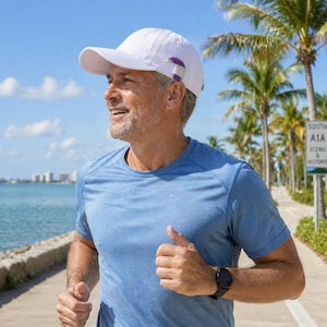 Peut inclure: Un homme portant une casquette de baseball blanche avec un clip violet, un t-shirt bleu et une montre noire, courant à l'extérieur. L'arrière-plan comprend des palmiers, un trottoir et un plan d'eau sous un ciel bleu.