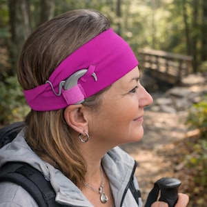 Puede incluir: Una mujer con una diadema magenta con un audífono adjunto, caminando al aire libre. La diadema asegura el audífono gris. Lleva una chaqueta gris y lleva una mochila y un bastón de senderismo.