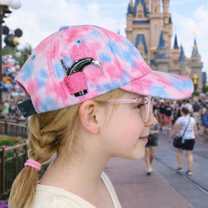 May include: A pink and blue tie-dye baseball cap featuring a small, black, curved hearing aid accessory attached to the side with a pink fabric band. The cap has a structured brim and a dark strap at the back, designed to hold a hearing device.