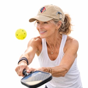 Może przedstawiać: Beżowa czapka z daszkiem z logo, noszona przez osobę grającą w pickleball. Osoba ta ma na sobie biały top na ramiączkach i trzyma rakietę do pickleballa. Żółta piłka do pickleballa jest w powietrzu.