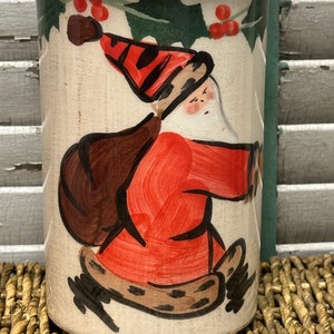 May include: A white ceramic mug with a hand-painted design of Santa Claus carrying a bag of gifts. The design includes holly leaves and red berries.