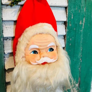 May include: A vintage Santa Claus head ornament with a red hat, white beard, and blue eyes. The ornament has a cheerful expression and is decorated with a sprig of holly. A festive decoration for the holiday season.