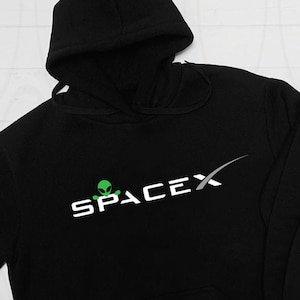 May include: Black hooded sweatshirt with a white graphic of the SpaceX logo and a green alien. The logo includes a stylized rocket trail.
