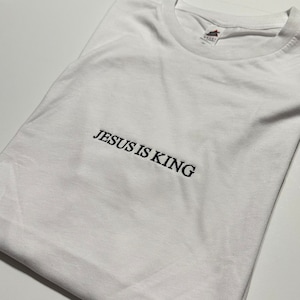 May include: White t-shirt with the embroidered text "JESUS IS KING" in black. The shirt is made of a soft material and has a crew neck. The shirt is folded and appears to be new.