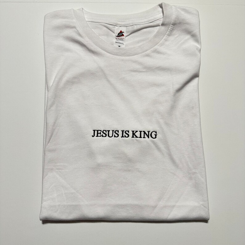 Puede incluir: Camiseta blanca con el texto "JESUS IS KING" impreso en negro en la parte delantera.