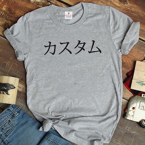 May include: A heather grey short-sleeved crew neck t-shirt featuring the black Japanese text "カスタム" printed across the chest. The shirt has a small white brand label at the inner neckline that reads "SMART BLANKS" with a small red logo icon.
