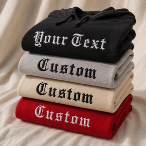 May include: A stack of four folded hooded sweatshirts in black, grey, beige, and red. The black hoodie features white embroidered text reading Your Text, while the grey, beige, and red hoodies feature black or white embroidered text reading Custom.
