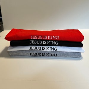 May include: A stack of four folded t-shirts in red, black, white, and gray. Each shirt has the embroidered text "JESUS IS KING" in white. The shirts are neatly stacked on a white surface.