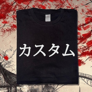 May include: A black short-sleeved t-shirt featuring white Japanese katakana text that reads "Kasutamu" printed across the centre of the chest. The garment has a standard crew neckline and a visible white brand label inside the collar area.