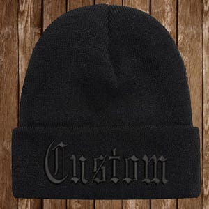 May include: A black beanie with the word "Custom" embroidered in a gothic font across the front. The beanie is made of a knitted material and is a solid black color.