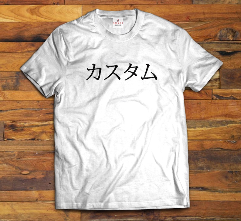 May include: A white short-sleeved t-shirt featuring the black Japanese text "カスタム" printed across the chest. The garment has a crew neckline and a small "SMART BLANKS" brand label located on the inside of the collar at the back of the neck.