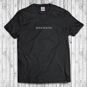 Puede incluir: Camiseta negra con el texto "JESUS IS KING" en letras doradas.