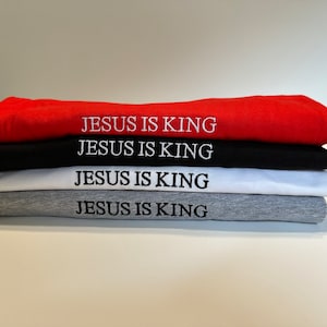 May include: A stack of four folded sweatshirts in red, black, white, and gray. Each sweatshirt has the embroidered text "JESUS IS KING" in white thread across the front.