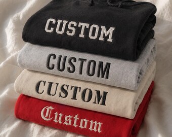 May include: A stack of four folded hooded sweatshirts in black, gray, beige, and red. Each hoodie features the word CUSTOM embroidered on the front. The top three hoodies use a block font, while the bottom red hoodie uses a gothic script font.