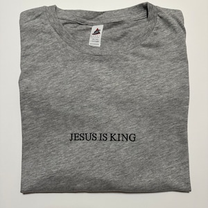 May include: A heather gray t-shirt with the embroidered text "JESUS IS KING" in black. The shirt is folded neatly, showcasing the crew neck and fabric texture. A tag is visible at the top.