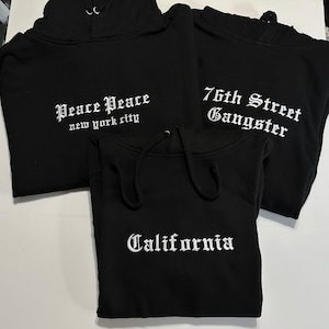 May include: Three black hooded sweatshirts with white embroidered text. The first sweatshirt says "Peace Peace new york city". The second sweatshirt says "76th Street Gangster". The third sweatshirt says "California".