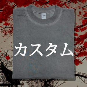 May include: A grey crew neck t-shirt featuring the white Japanese text "カスタム" printed across the centre of the chest. The garment has a ribbed collar and a small white brand label visible on the inside of the neckline, showcasing a simple design.
