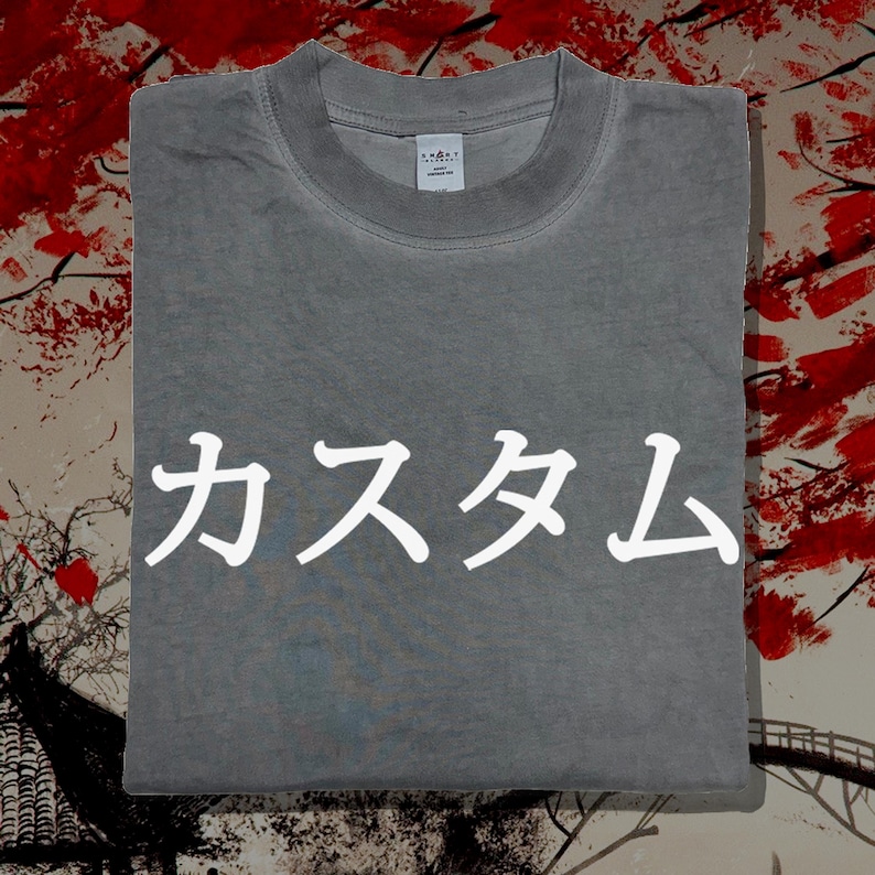 May include: A grey crew neck t-shirt featuring the white Japanese text "カスタム" printed across the centre of the chest. The garment has a ribbed collar and a small white brand label visible on the inside of the neckline, showcasing a simple design.