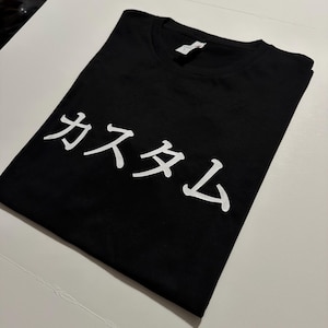 May include: A black short-sleeved t-shirt featuring white Japanese katakana text that reads "kasutamu" printed across the chest. The garment has a crew neckline and a standard fit, showcasing a clean design with high-contrast white lettering.