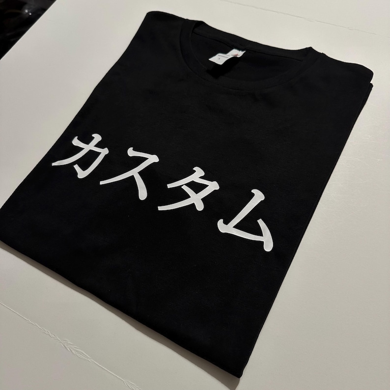 May include: A black short-sleeved t-shirt featuring white Japanese katakana text that reads "kasutamu" printed across the chest. The garment has a crew neckline and a standard fit, showcasing a clean design with high-contrast white lettering.