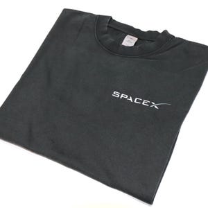 May include: A dark gray t-shirt with the word "SPACEX" printed in white on the front. The shirt is folded and appears to be made of a soft material. The t-shirt has a crew neck.