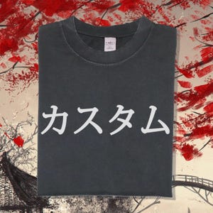May include: A charcoal grey short-sleeved t-shirt featuring white Japanese text that reads "カスタム" printed across the centre of the chest. The garment has a crew neckline and a visible white brand label inside the collar area.