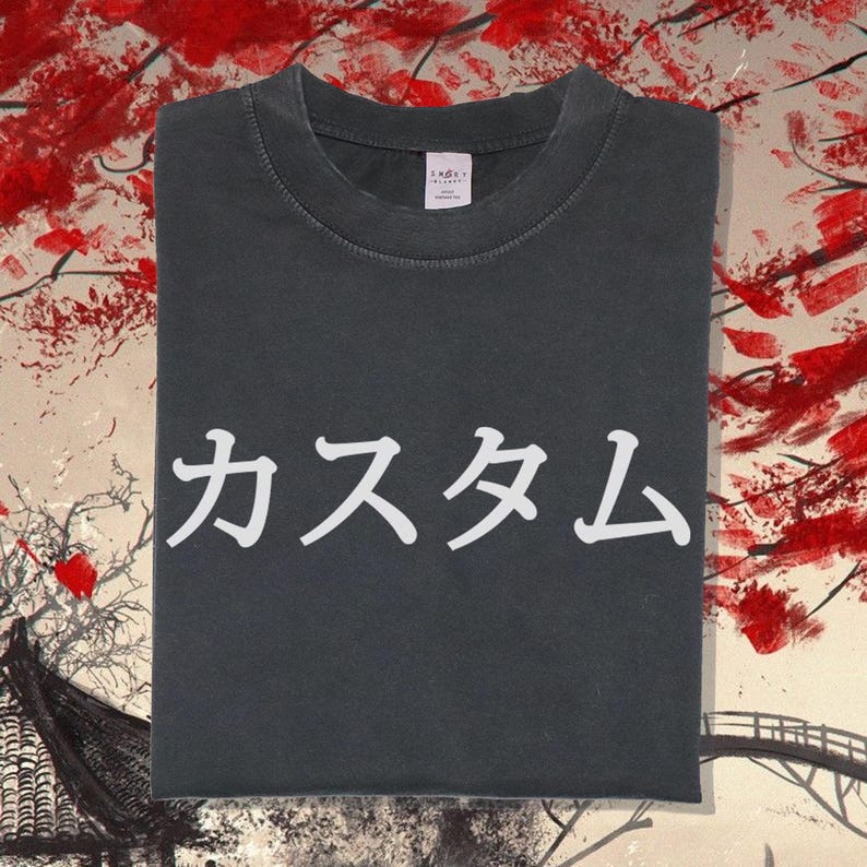 May include: A charcoal grey short-sleeved t-shirt featuring white Japanese text that reads "カスタム" printed across the centre of the chest. The garment has a crew neckline and a visible white brand label inside the collar area.