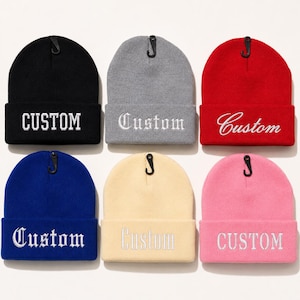 May include: Six knit beanies in black, gray, red, blue, cream, and pink, each featuring a cuffed brim and white embroidered text reading Custom in various fonts. Each beanie includes a small black plastic hanging hook at the top center.