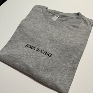 Puede incluir: Una camiseta gris con el texto "JESUS IS KING" bordado en negro.