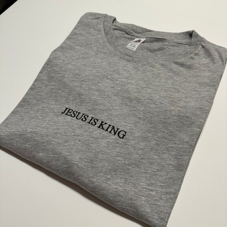 Puede incluir: Una camiseta gris con el texto "JESUS IS KING" bordado en negro.