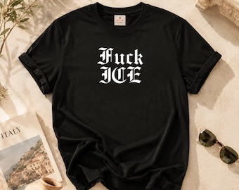 May include: A black short-sleeved t-shirt featuring the white text "FUCK ICE" printed in a bold, gothic-style font across the center of the chest. The garment has a standard crew neckline and a small brand label visible inside the collar area.