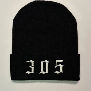 May include: Black knit beanie with the white embroidered number "305" in a gothic font. The beanie is folded at the bottom and is made of a soft, warm material.