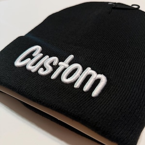 May include: A black beanie with the word "Custom" embroidered in white script across the front. The beanie is made of a knitted material and has a folded brim.