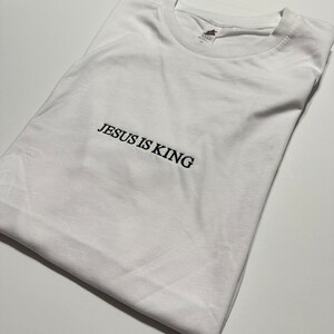 Puede incluir: Camiseta blanca con el texto "JESUS IS KING" bordado en negro.