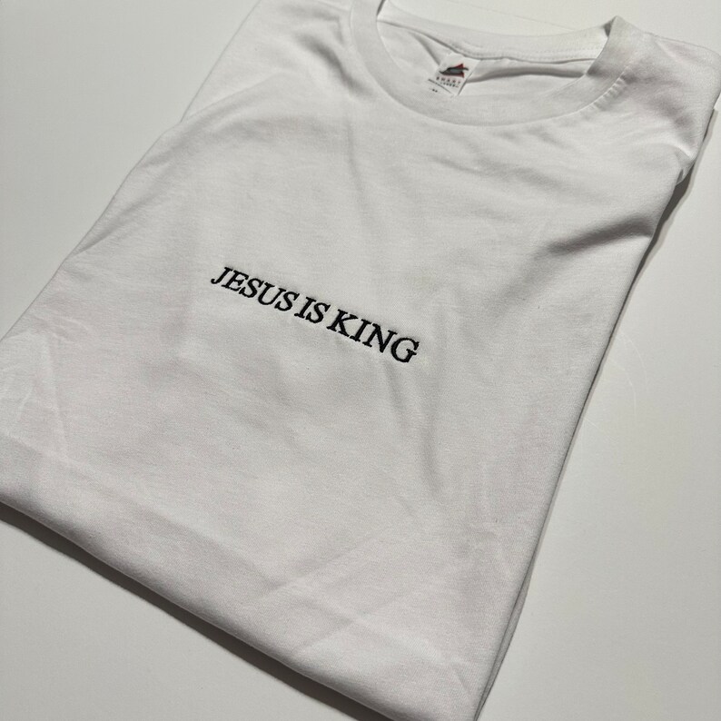Puede incluir: Camiseta blanca con el texto "JESUS IS KING" bordado en negro.