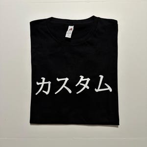 May include: A black short-sleeved t-shirt featuring the white Japanese text "カスタム" printed across the centre of the chest. The garment has a crew neckline and a white interior brand label attached to the collar, indicating a size large.
