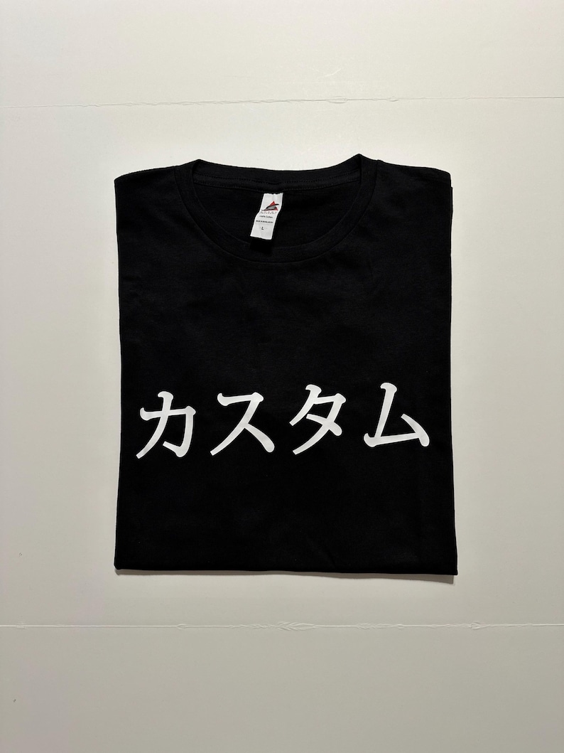 May include: A black short-sleeved t-shirt featuring the white Japanese text "カスタム" printed across the centre of the chest. The garment has a crew neckline and a white interior brand label attached to the collar, indicating a size large.