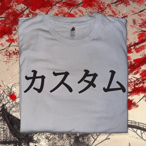 May include: A white short-sleeved t-shirt featuring black Japanese text printed across the centre of the chest. The shirt has a crew neckline and a visible brand label inside the collar. The fabric is plain white cotton with a standard fit.