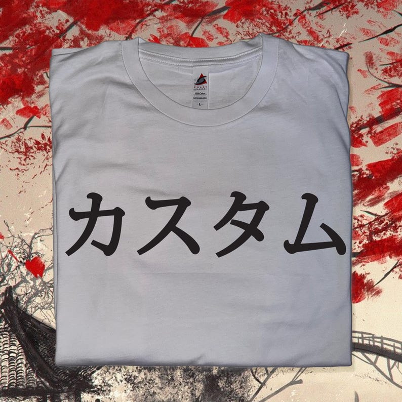 May include: A white short-sleeved t-shirt featuring black Japanese text printed across the centre of the chest. The shirt has a crew neckline and a visible brand label inside the collar. The fabric is plain white cotton with a standard fit.
