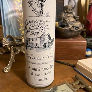 May include: A white candle with a black and white illustration of a house and a woman holding a rake. The text on the candle reads "Beautiful Nest", "Bellissimo Nido", "A ogni uccello il suo nido &egrave; bello."