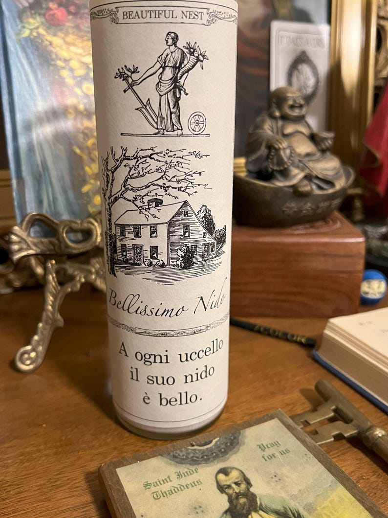 May include: A white candle with a black and white illustration of a house and a woman holding a rake. The text on the candle reads "Beautiful Nest", "Bellissimo Nido", "A ogni uccello il suo nido &egrave; bello."