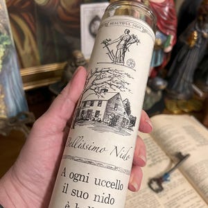 May include: A white candle with a black and white illustration of a house and a woman holding a branch with leaves. The text on the candle reads "Bellissimo Nido" and "A ogni uccello il suo nido &egrave; bello."