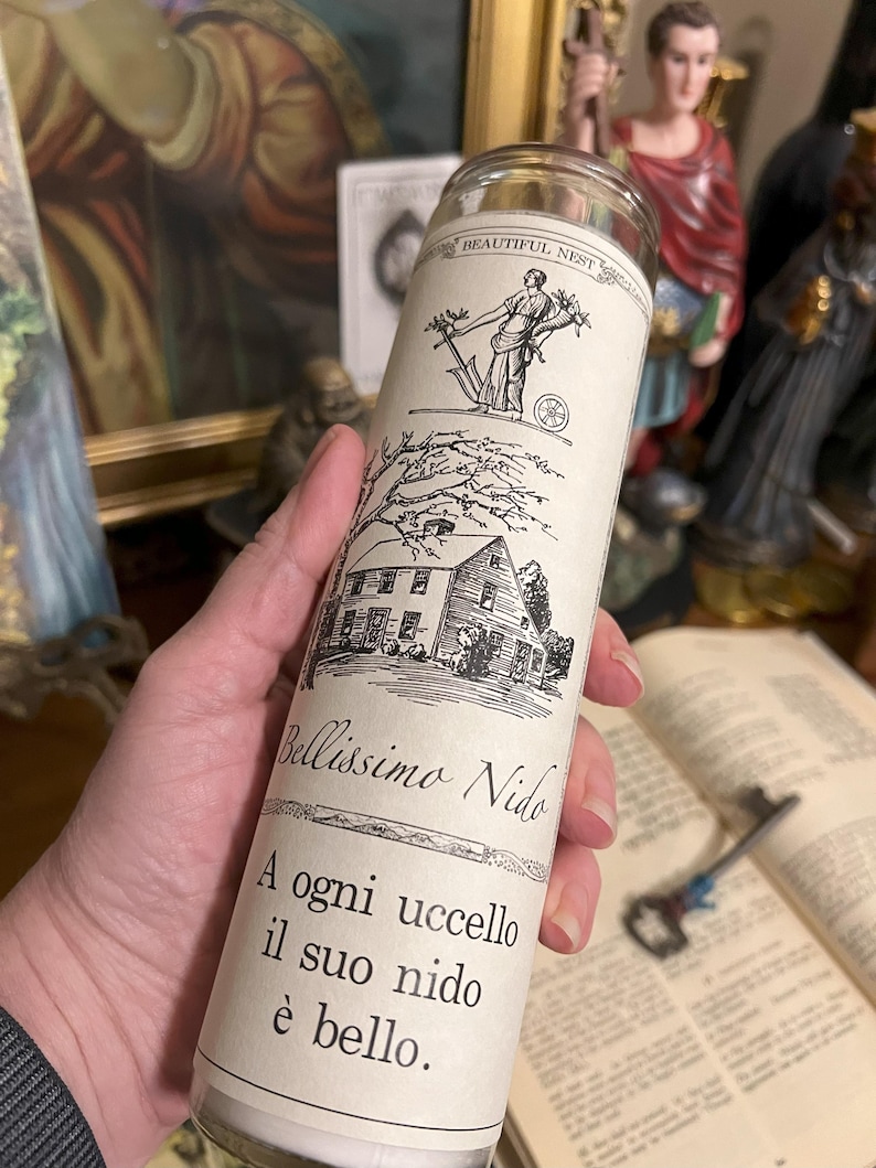 May include: A white candle with a black and white illustration of a house and a woman holding a branch with leaves. The text on the candle reads "Bellissimo Nido" and "A ogni uccello il suo nido &egrave; bello."