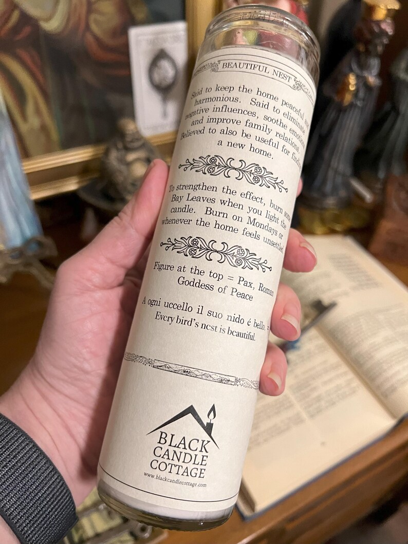 May include: White candle with a black and white label. The label says "Beautiful Nest" and includes text about the candle's use and the Roman Goddess of Peace. The bottom of the label says "Black Candle Cottage" and includes a website address.