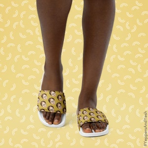 May include: Yellow slide sandals featuring a repeating pattern of a man's face. The sandals have a white sole and a wide strap across the top of the foot. The background is a light yellow color with a repeating pattern of white curved lines.