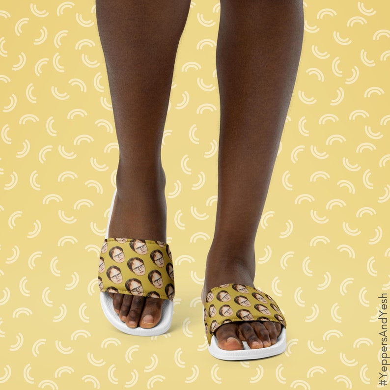 May include: Yellow slide sandals featuring a repeating pattern of a man's face. The sandals have a white sole and a wide strap across the top of the foot. The background is a light yellow color with a repeating pattern of white curved lines.